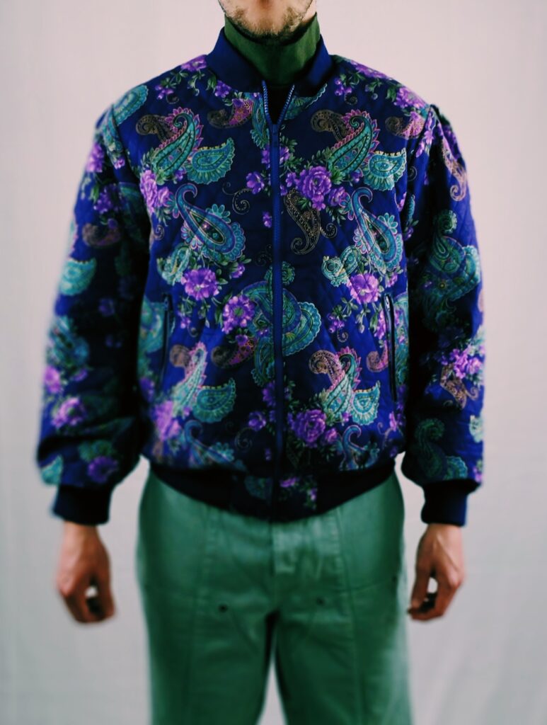 Purple floral/paisley bomber jacket. The model is styled in a green Sweater by top man, green jeans by jacquemus, brown belt by velez and brown shoes.