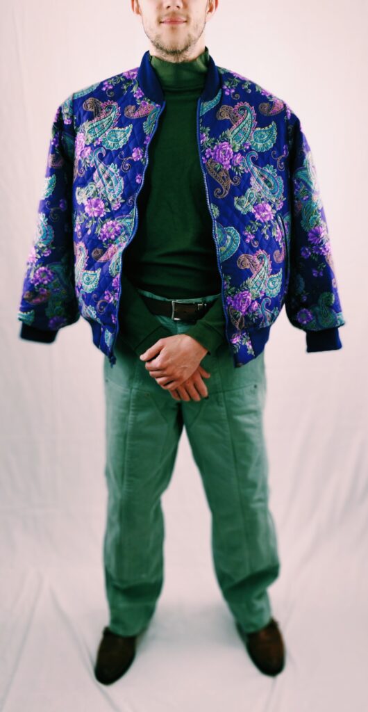 Purple floral/paisley bomber jacket. Styled with a green sweater and green jeans, brown belt and shoes.