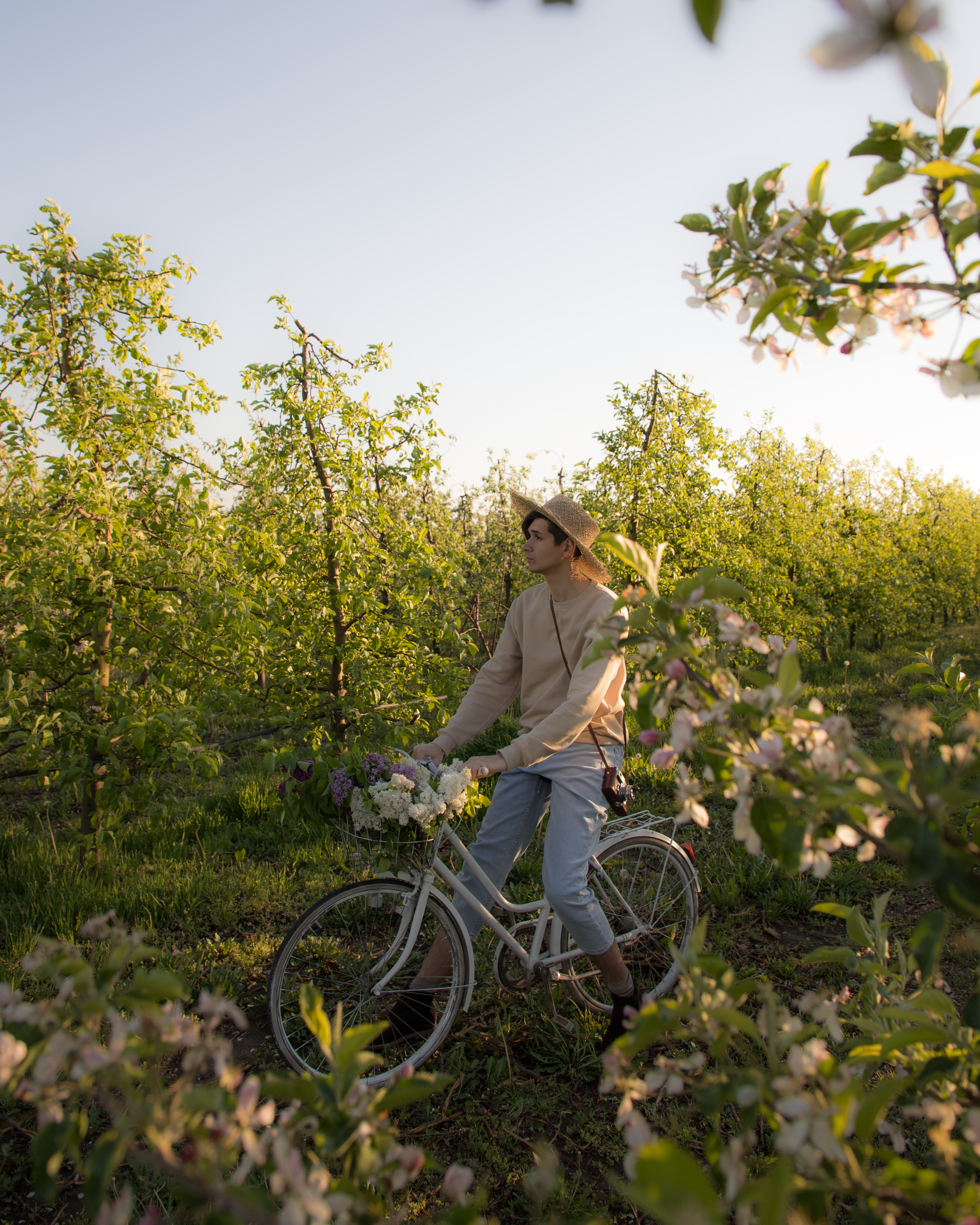 A guy on a forest wearing crop pants, boots, a straw hat and a sweater. He is on a bike with a basket on the front. The basket is full of flowers. There are also flowers around him. The style is Cottagecore.