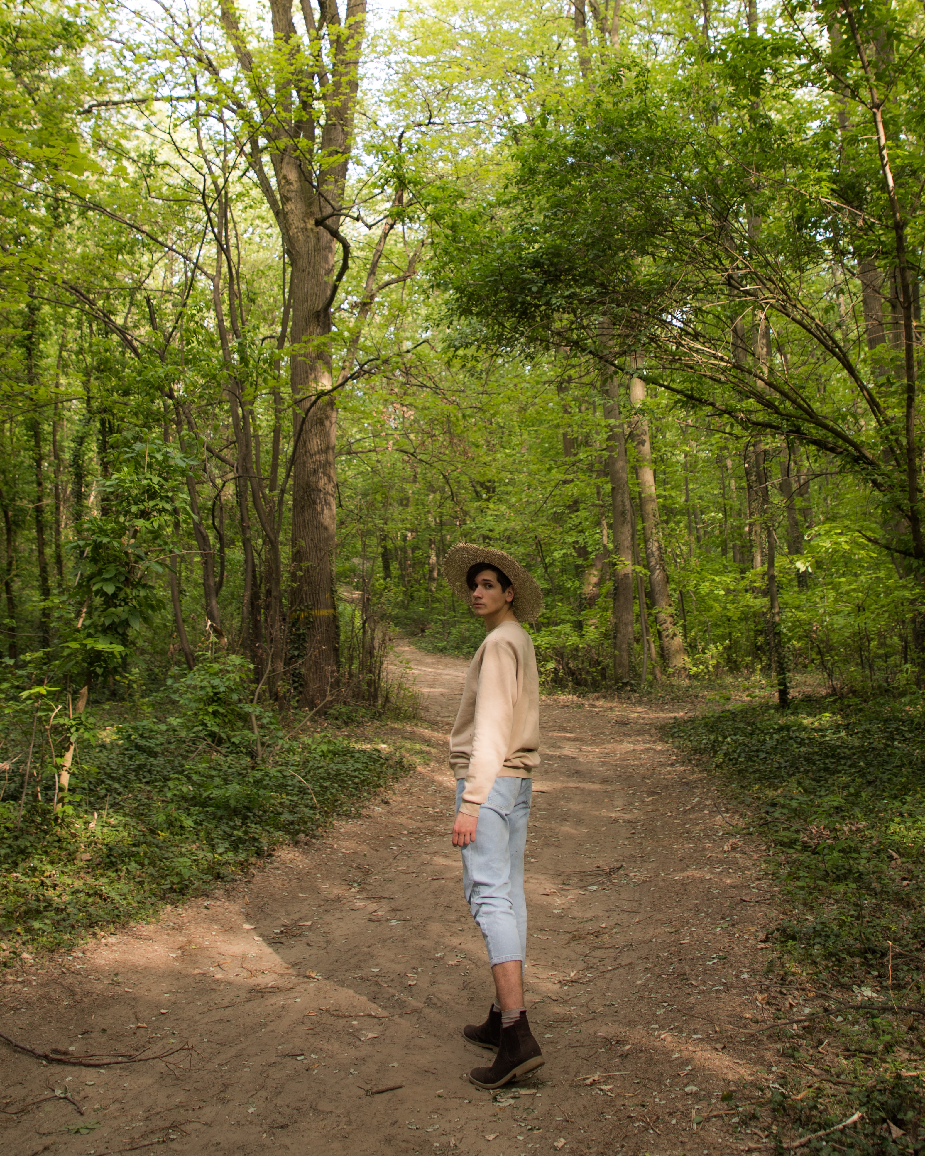 A guy on a forest wearing crop pants, boots, a straw hat and a sweater. The style is Cottagecore.